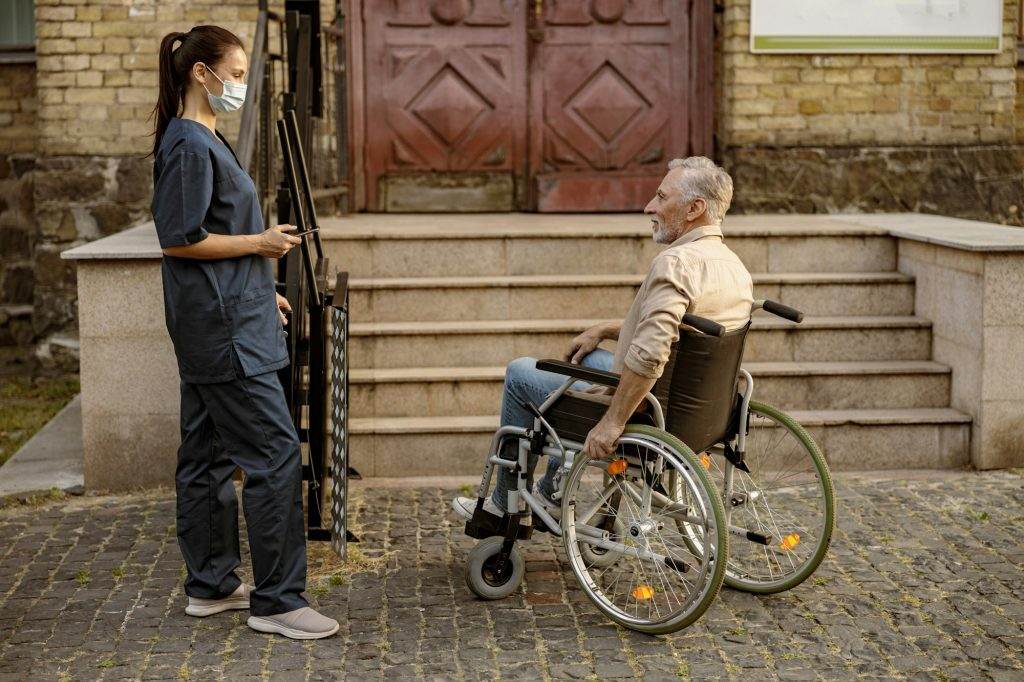 Full length shot of lovely young nurse in protective face mask helping mature disabled man in