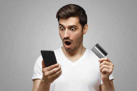 Portrait of shocked man, holding credit card and phone, having no money or blocked bank account