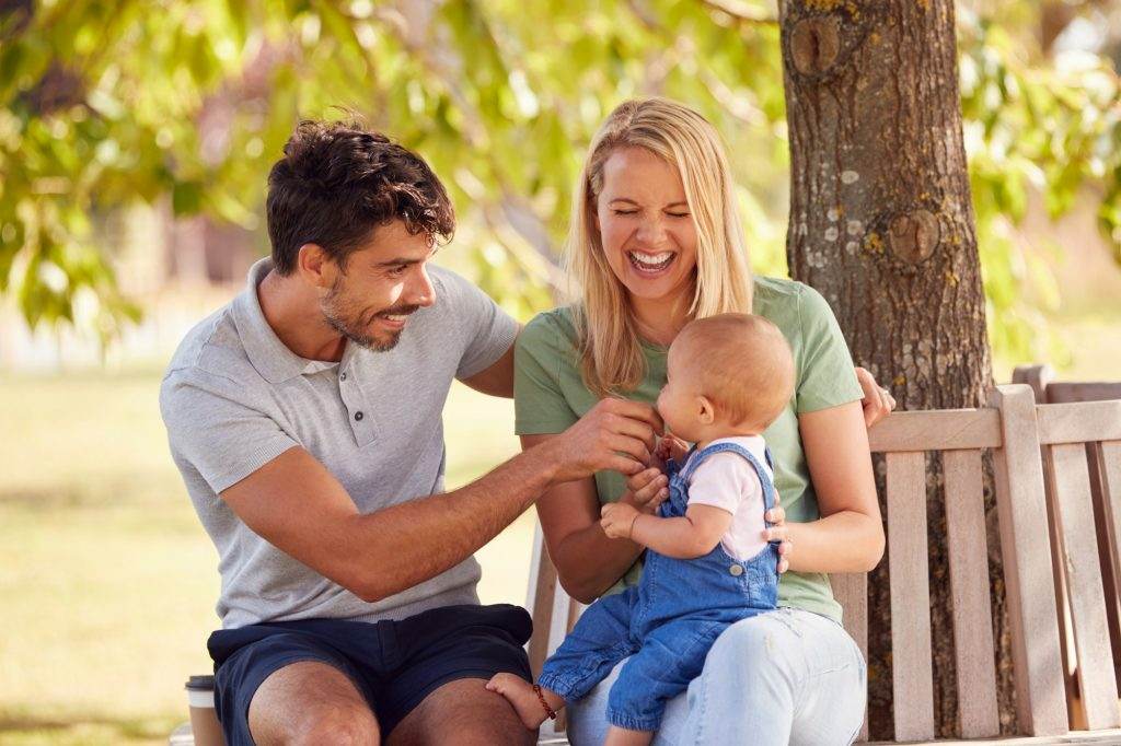 Family With Baby Daughter Sitting On Seat Under Tree In Summer Park Together