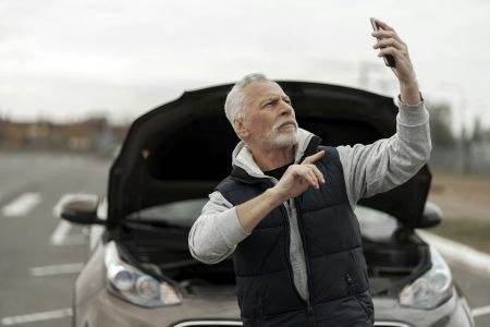 Elderly man driver talking on smartphone near broken auto with open hood. Car insurance concept