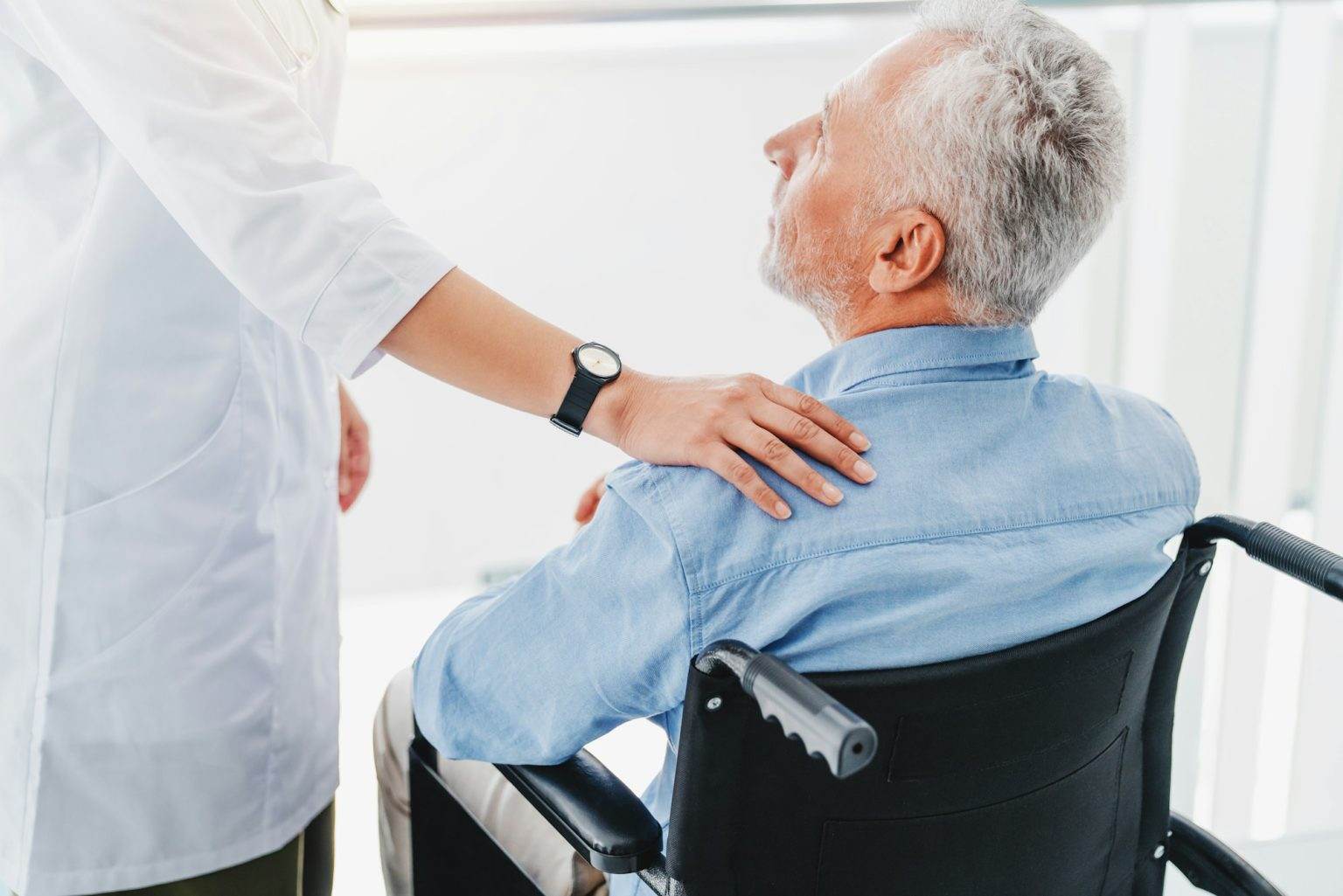 Female doctor supporting unhappy and unhealthy handicapped patient in wheelchair indoors.
