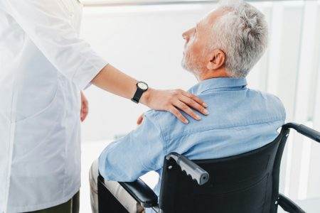 Female doctor supporting unhappy and unhealthy handicapped patient in wheelchair indoors.