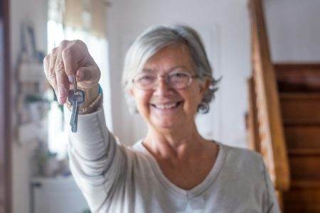 Close up of old and mature woman at home holding keys of house to the camera.