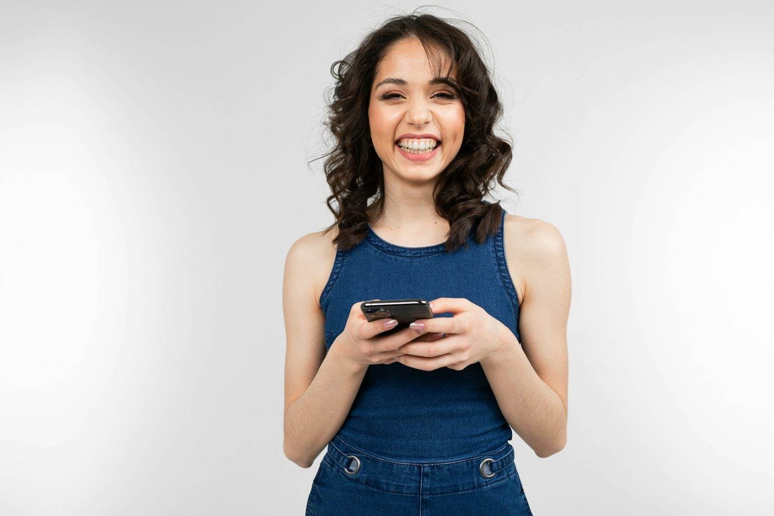joyful girl with a smile in a blue dress holds a smartphone and glitters with happiness on a white