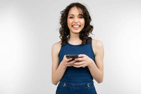 joyful girl with a smile in a blue dress holds a smartphone and glitters with happiness on a white