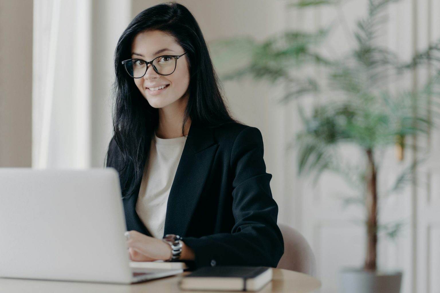 Smiling well dressed young woman secretary with dark hair, works on laptop computer at desktop