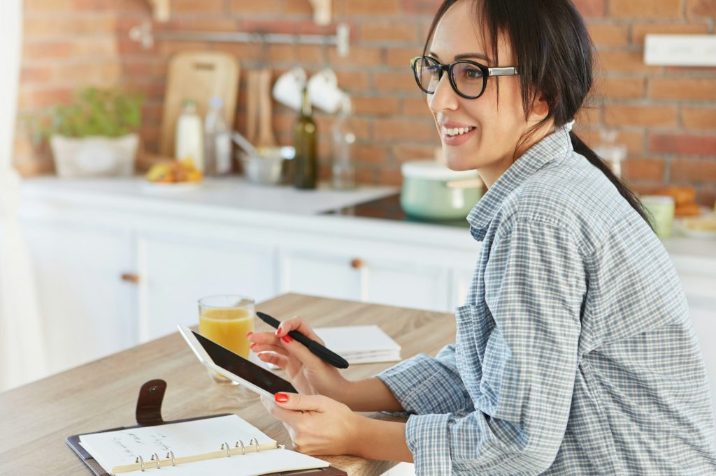 Portrait of cheerful brunette woman wears casual shirt, makes menu for birthday party, uses modern t