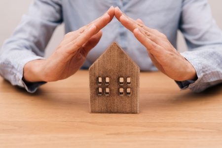 close up of Hands forming roof over wooden house model on wooden desk, home insurance concept