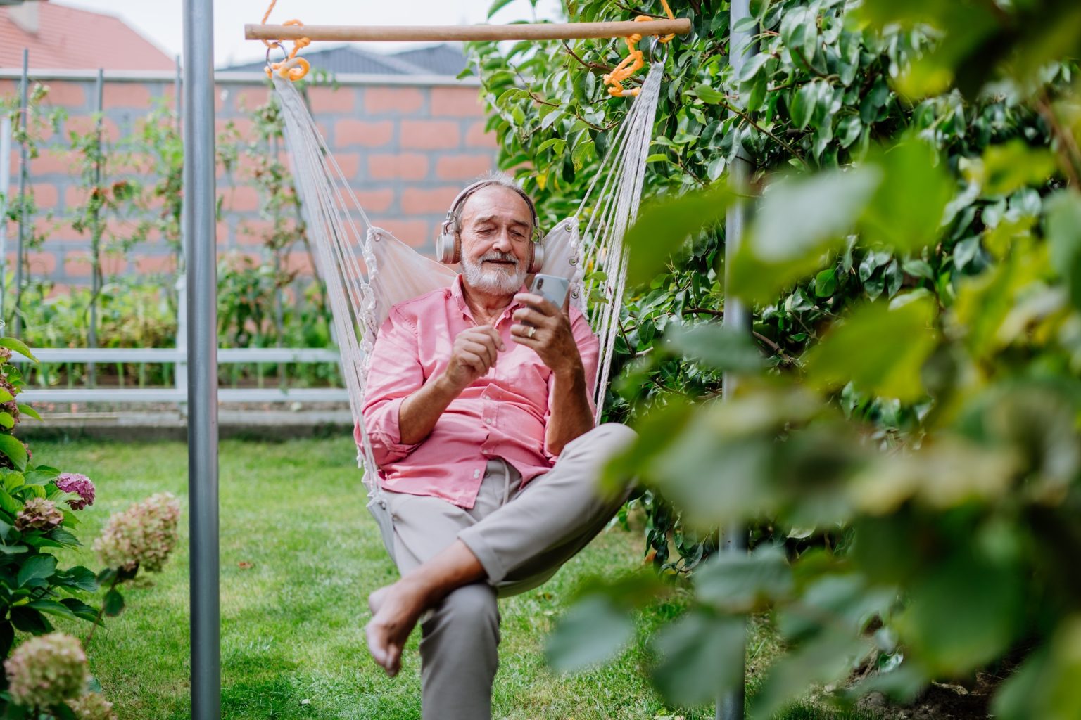 Happy senior man sitting in outdoor garden swing, listening music in headphones and scrolling