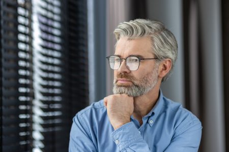 Thoughtful mature man with glasses reflecting by window in well-lit room