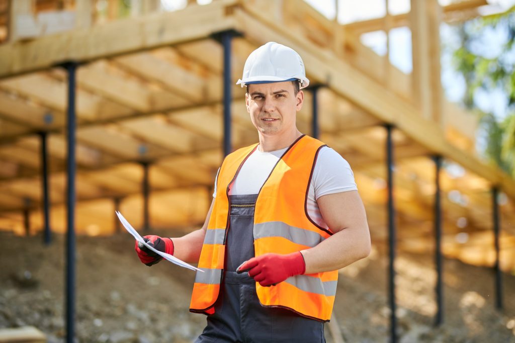 Young man wearing yellow vest, uniform and white helmet, standing, building house.