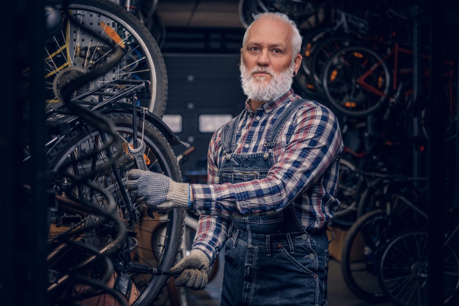Elderly handyman dressed in plaid shirt and overalls around bikes