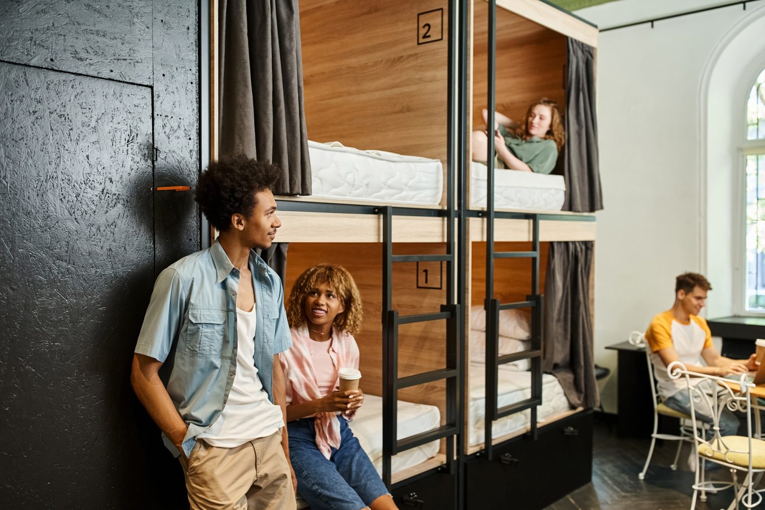 happy african american woman with coffee to go talking to friend near roommates in cozy hostel room