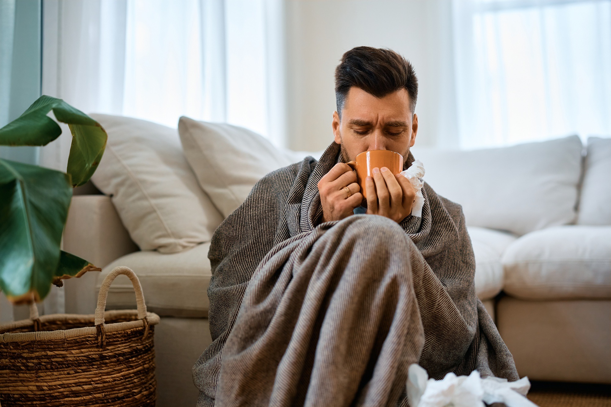 Sick man wrapped in blanket drinking tea at home.