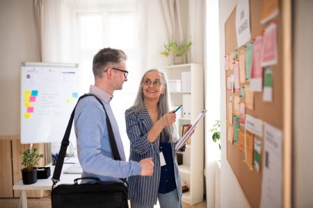 Recruitment agency employee standing in front of employment noticeboard and helping mature man