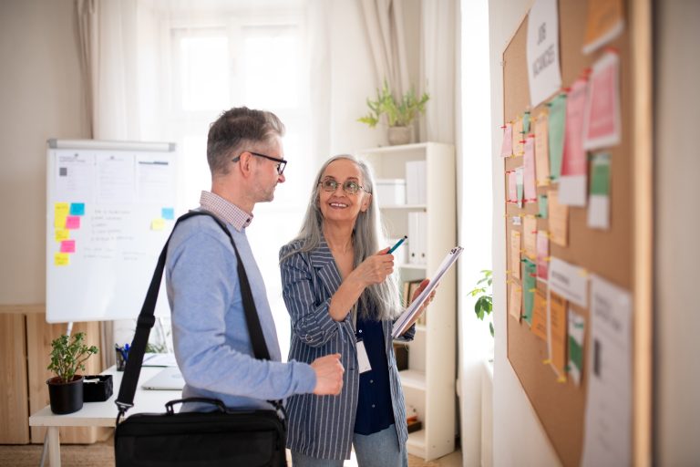 Recruitment agency employee standing in front of employment noticeboard and helping mature man
