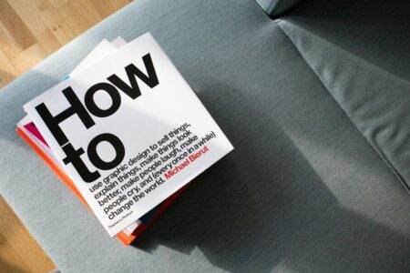 Comment choisir le meilleur super livret bancaire en 2026 A collection of design books on a couch, showcasing 'How to' by Michael Bierut