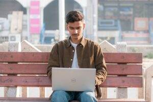 A young man working on his laptop while sitting on an outdoor bench in a city environment.