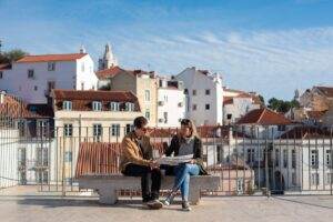 L’état des lieux locatif : les 7 pièges qui coûtent 500 € en moyenne au dépôt de garantie A couple sitting on a bench looking at a map with Lisbon's rooftops in the background.