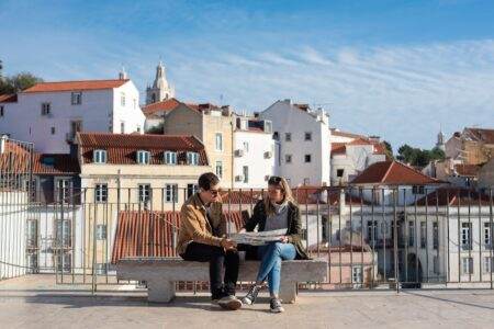 L’état des lieux locatif : les 7 pièges qui coûtent 500 € en moyenne au dépôt de garantie A couple sitting on a bench looking at a map with Lisbon's rooftops in the background.