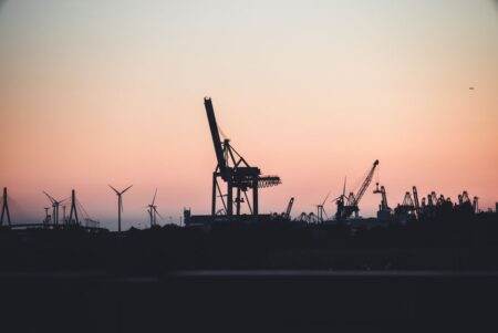 Silhouetted cranes and wind turbines at the port of Hamburg during sunset, highlighting industrial beauty.