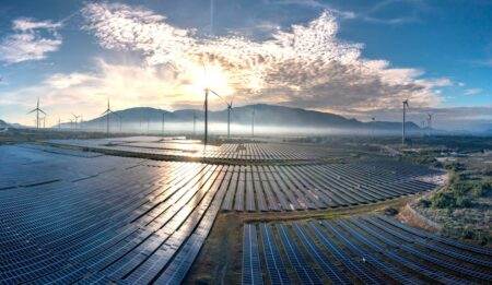 Grève EDF : pourquoi l’arrêt de plusieurs réacteurs nucléaires ne fait pas (encore) vaciller le système électrique Stunning aerial view of a vast solar farm with wind turbines under a vibrant sunrise sky, showcasing renewable energy.