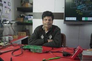 An electronics engineer works at the repair lab desk, surrounded by tools and a circuit board.