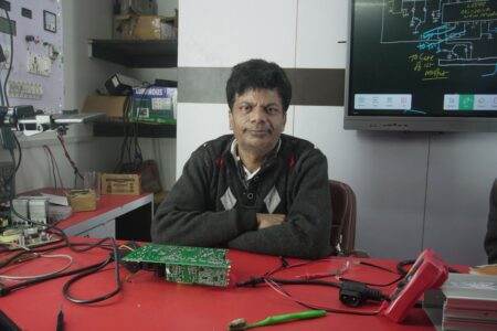 An electronics engineer works at the repair lab desk, surrounded by tools and a circuit board.