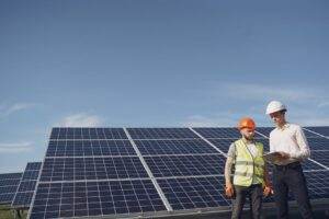 From below of serious young male colleagues in helmets working at solar energy station against cloudless blue sky