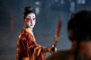 A captivating geisha in costume poses dramatically with a weapon in hand onstage.