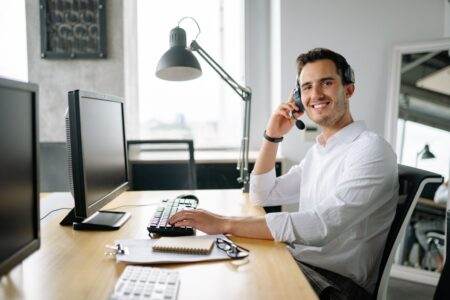 Services d’assistance pannes JeChange : ce qu’il faut savoir avant de souscrire Smiling call center agent working at a desk with a headset and computer in a modern office.