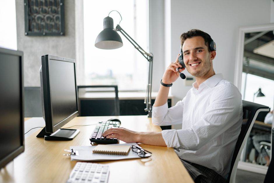 Services d’assistance pannes JeChange : ce qu’il faut savoir avant de souscrire Smiling call center agent working at a desk with a headset and computer in a modern office.