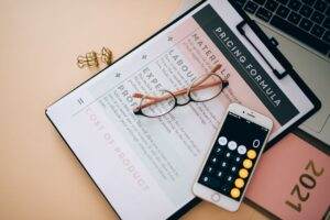 Overhead view of business tools including a phone calculator, pricing formula document, and eyeglasses on a desk.