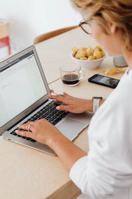 Adult woman working from home, typing on laptop with coffee and snacks on wooden table.
