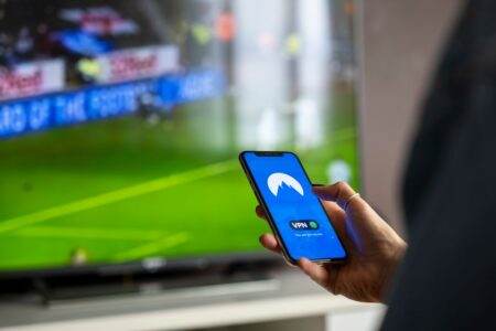 Proton VPN Free ou Plus : prix, limites et usages en 2026 Close-up of a person holding a smartphone with a VPN app, streaming sports on TV.