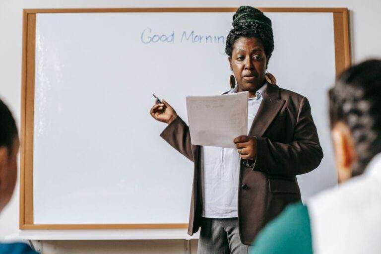 Rentrée scolaire 2026 : le cartable ne suffit pas Concentrated adult African American female teacher wearing formal suit standing near whiteboard and talking to pupils in light classroom