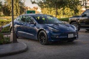 Electric blue Tesla Model 3 parked in sunlit commercial parking area.