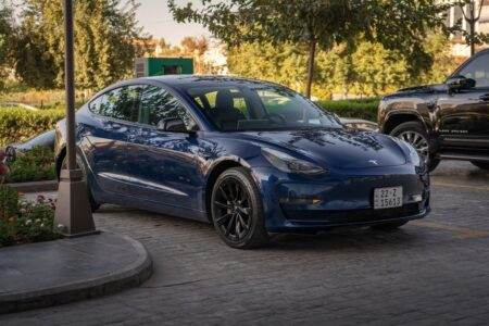 Electric blue Tesla Model 3 parked in sunlit commercial parking area.