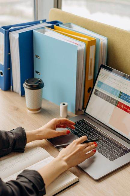 Hands typing on a laptop at an office desk with files and coffee.