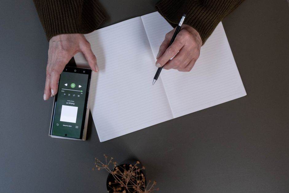Top view of a person writing on paper with smartphone displaying music app.