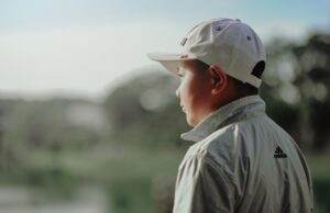 A boy wearing a white cap and jacket looks thoughtfully into the distance outdoors.