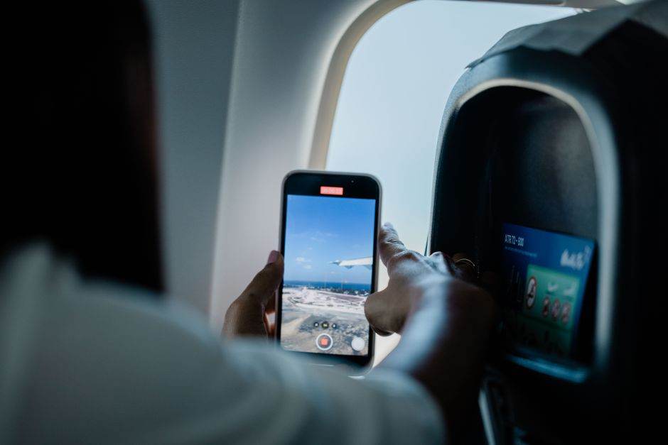 A passenger films the outside view from an airplane window using their smartphone, capturing the wing and sky.