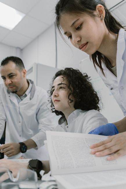 Three scientists wearing lab coats collaborate on research with books and computers in a modern laboratory setting.