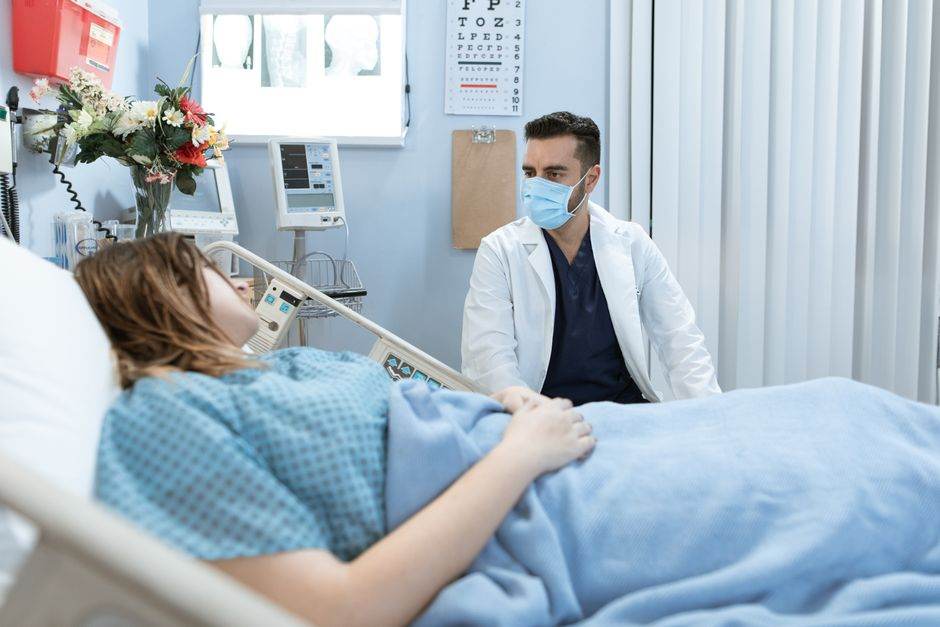 A doctor in a face mask talks to a patient in a hospital bed, providing care and consultation.
