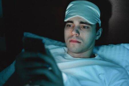 Applications de sommeil en 2026 : ce que mesurent les scores A man wearing a sleeping mask uses his smartphone while lying in bed at night.