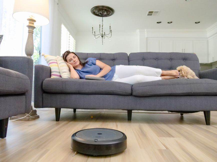 A woman relaxing on a sofa with a robotic vacuum cleaner cleaning the hardwood floor.