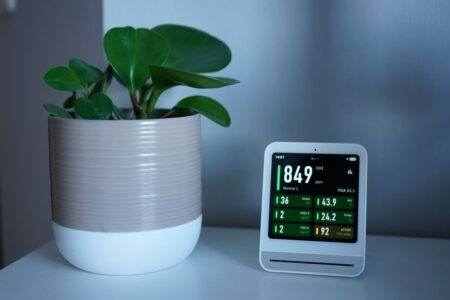 Purificateur d’air dans une chambre : ce qu’il filtre et ses limites Air quality monitor next to a potted plant on a desk, showing CO2 and PM2.5 levels.